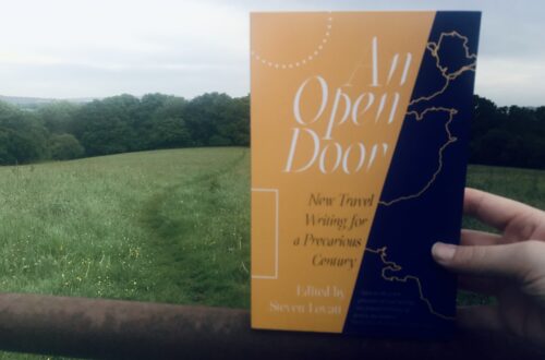 A copy of An Open Door anthology is held up on a rusty gate, a green meadow seen behind with a path through the grass, trees beyond.