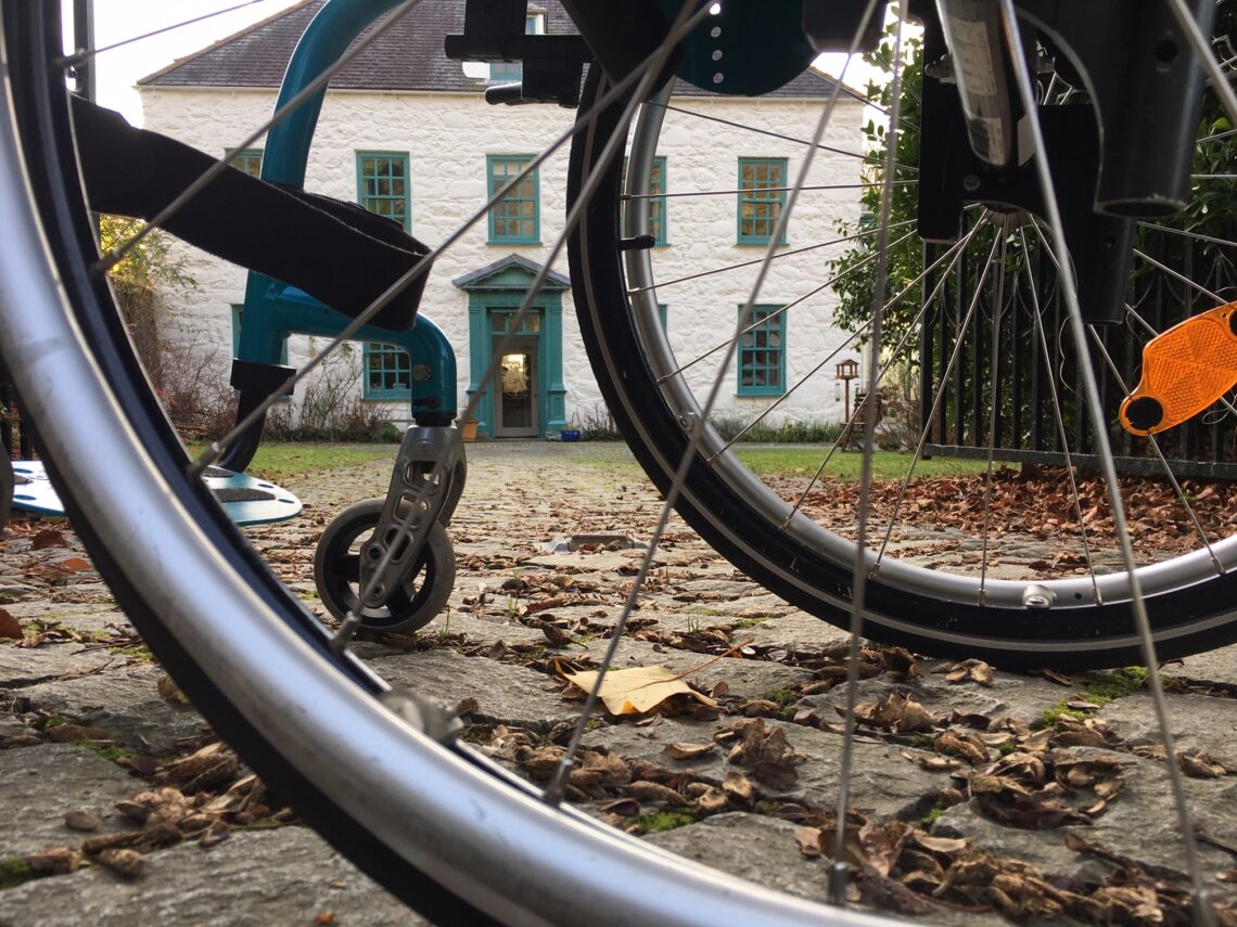 Large white house with teal door seen through Grace's wheelchair wheels. Leaves on the ground under the chair.