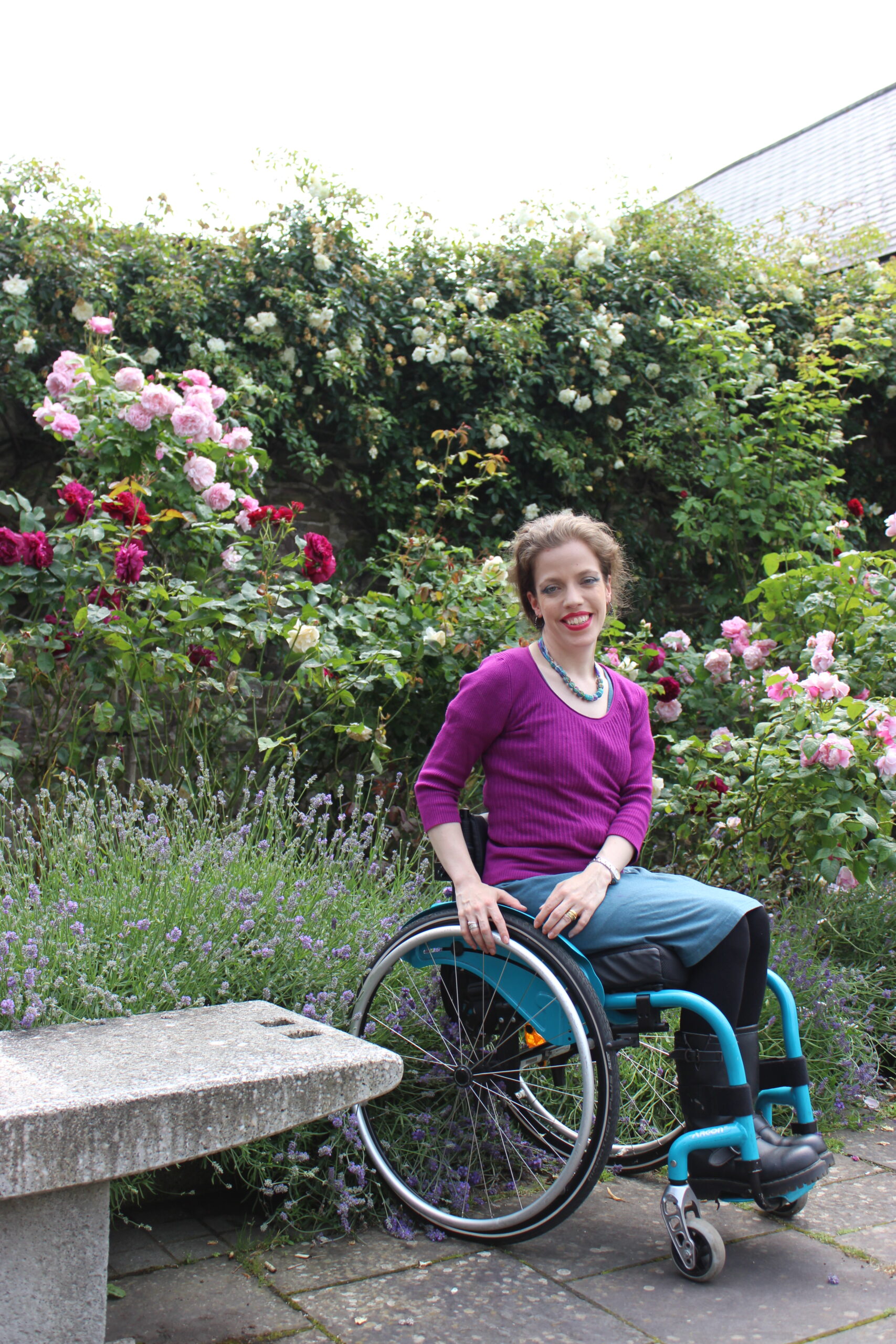 Grace sitting in her teal wheelchair in front of roses in bloom wearing a pink jumper and a teal skirt.
